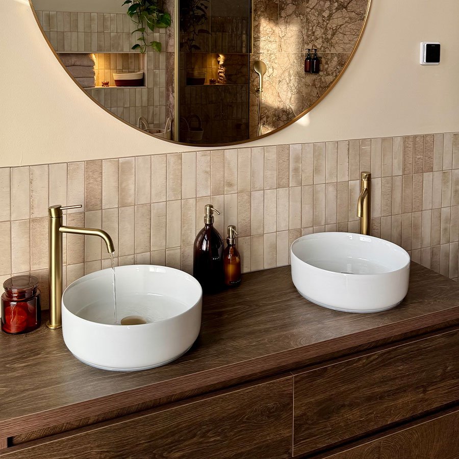 Donker houten badkamermeubel met beige wandtegels, witte waskommen, gouden kranen en een grote ronde spiegel