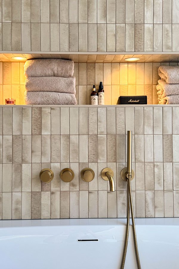 Beige wandtegel met nis met spotjes erin boven het bad met een gouden badkraan