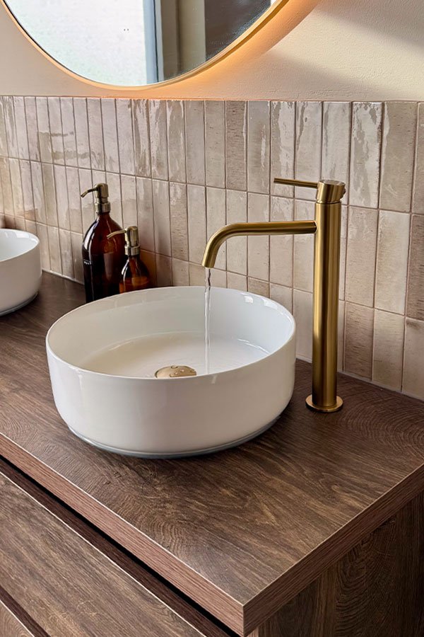 Beige wandtegel, gouden kraan en een witte waskom op een donker houten meubel